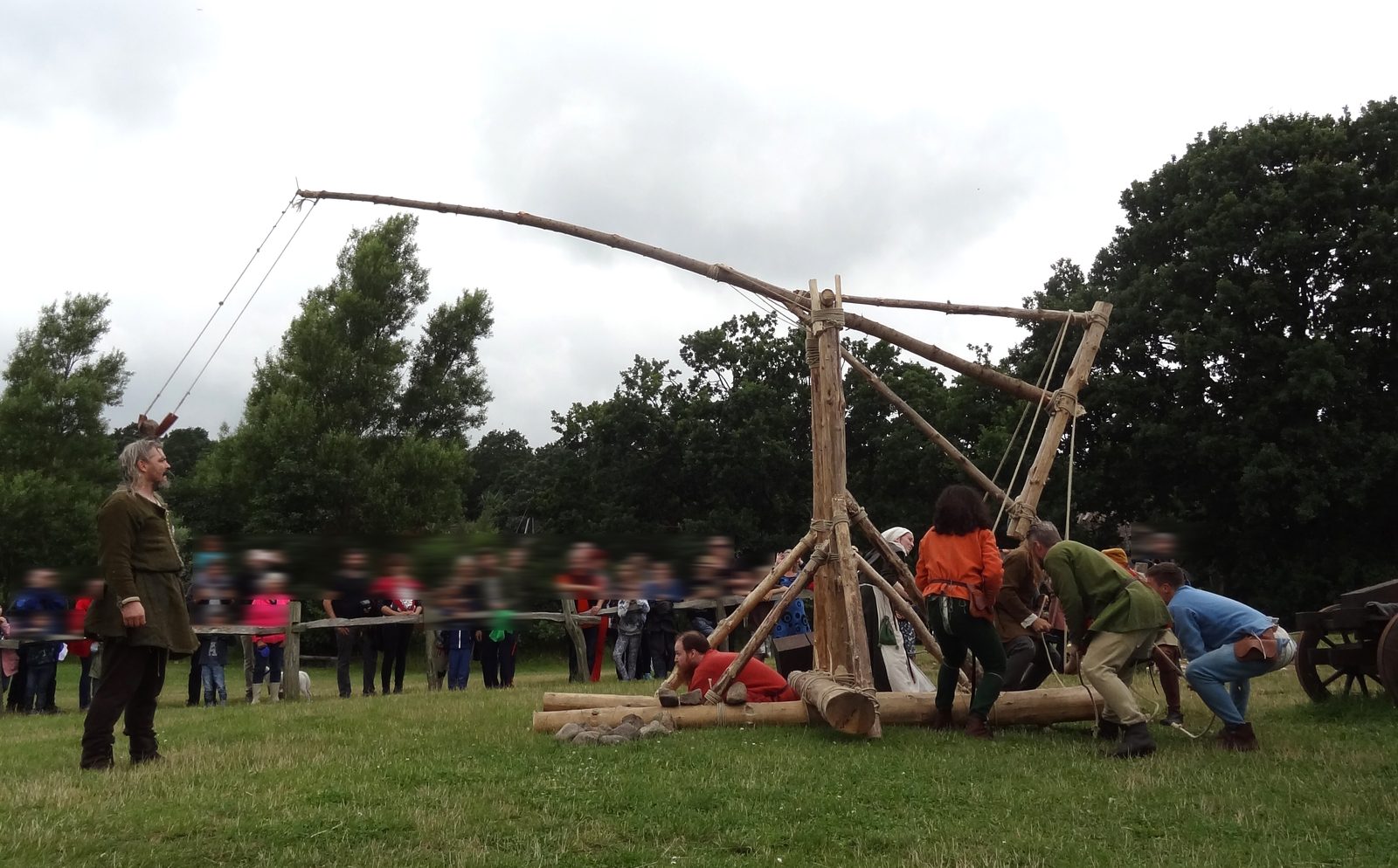 A traction trebuchet built from unmilled logs on a festival field, its throwing arm swung up and over after release, sling ropes trailing. The crew stands at the machine and spectators watch from behind a rope line to the side.