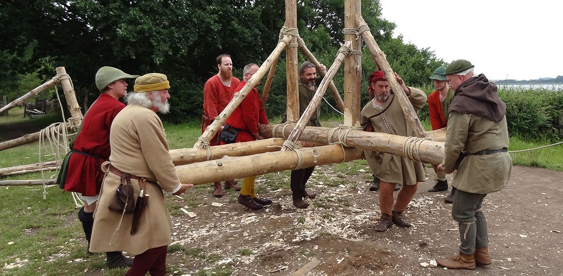 Nine people in medieval dress lift an assembled trebuchet frame of lashed logs waist-high and carry it across the build site, with woodchips from the construction still on the ground.